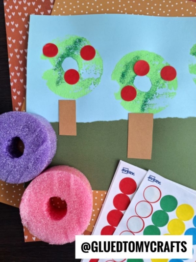 Children’s apple tree craft made with a blue and green paper background, sponge-painted green tree tops, brown trunks, and red dot stickers as apples. Perfect for pool noodle crafts—two cut pool noodles and colored dot stickers are nearby.