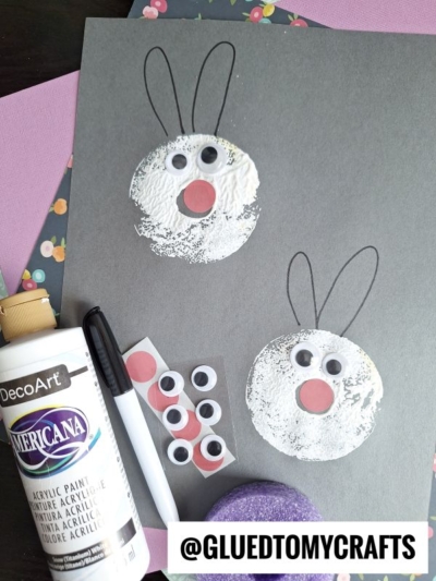 A gray paper showcases two bunny faces made from white paint circles, with black marker ears, googly eyes, and pink noses—an adorable idea to try with pool noodle crafts. Supplies like paint, a sponge, and craft papers surround the artwork. @GLUEDTOMYCRAFTS is at the bottom.