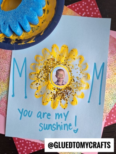 A handmade Mother’s Day card reads MOM, you are my sunshine! with a yellow painted flower, a child’s photo in the center, and colorful paper layers behind. Blue paint, a sponge, and pieces from a pool noodle craft are visible beside the card.