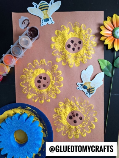 A brown paper features three painted yellow sunflowers with brown centers and black dots, two bee stickers, a plastic flower from a pool noodle craft, a blue sponge, and paint with brushes. @GLUEDTOMYCRAFTS is written at the bottom.
