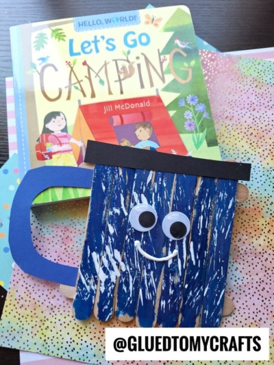 A childrens book titled Lets Go Camping lies on a colorful background next to a blue popsicle stick camping mug with googly eyes and a smile. The mug’s handle is made from paper. @GLUEDTOMYCRAFTS is at the bottom.