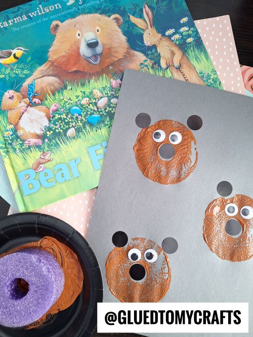 Children’s bear-themed craft with brown paint-stamped bear faces on gray paper, created using a pool noodle craft technique and googly eyes, next to the book Bear Finds. ©GLUEDTOMYCRAFTS.