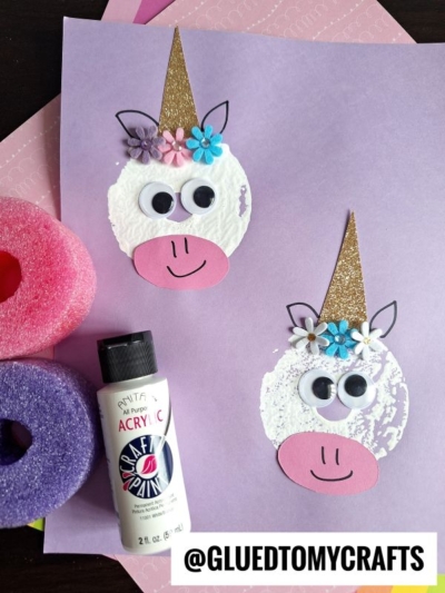 Two unicorn faces made from white paint circles and paper details are on purple paper. Each has googly eyes, pink snouts, glittery gold horns, and paper flowers—a magical twist perfect for pool noodle crafts. @GLUEDTOMYCRAFTS is written.