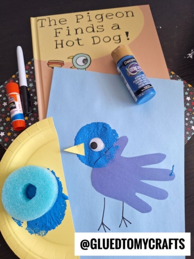 A children’s craft project inspired by The Pigeon Finds a Hot Dog! features a blue handprint bird with a googly eye on blue paper, next to pool noodle crafts supplies and the book. The Instagram handle @GLUEDTOMYCRAFTS is at the bottom.