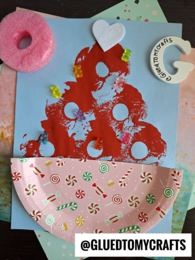 A kids’ craft project featuring paper plate candy, a painted red tree with circular stamps, foam heart, candy-shaped beads, a pink donut shape, and a puzzle piece with “GluedToMyCrafts”—plus fun pool noodle crafts touches! @GLUEDTOMYCRAFTS label at the bottom.