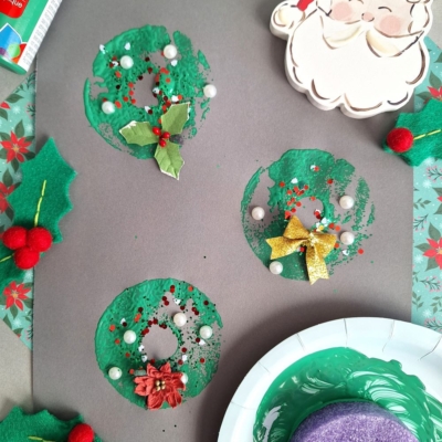 A festive Christmas craft project featuring green painted wreaths made from pool noodle crafts with glitter, bows, and pom-poms on gray paper, surrounded by holly decorations, paint supplies, and a smiling Santa cutout. @GLUEDTOMYCRAFTS appears at the bottom.