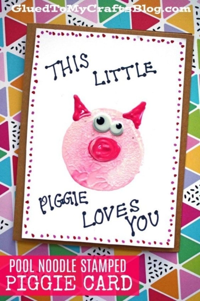 A handmade card featuring a cute pink pig face from a pool noodle craft, with googly eyes and pink details. The card says "This little piggie loves you" and sits on a vibrant geometric background.