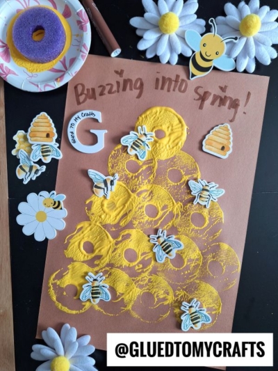 A brown paper with yellow paint stamped in circles—using a pool noodle craft technique—bee and flower stickers, and “Buzzing into Spring!” on top. Craft supplies, a purple sponge, daisies, and @GLUEDTOMYCRAFTS appear at the bottom.