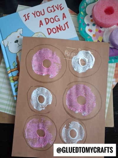 A brown sheet with six donut shapes—made using a pool noodle craft and decorated in pink and white paint—sits atop the children’s book “If You Give a Dog a Donut.” Craft supplies are nearby. @GLUEDTOMYCRAFTS watermark is visible.