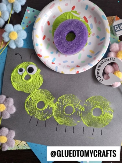 A caterpillar is stamped onto black paper using green paint and a pool noodle craft donut shape. Googly eyes and drawn antennae complete the look. Craft supplies, flowers, and @GLUEDTOMYCRAFTS watermark surround the artwork.