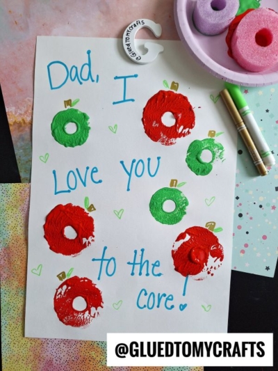 A handmade card for Dad says “Dad, I love you to the core!” with red and green apple prints made from stamped pool noodle crafts. Markers and art supplies are nearby. @GLUEDTOMYCRAFTS is written at the bottom.