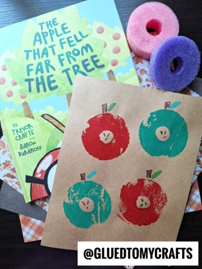 A children’s book titled The Apple That Fell Far From the Tree, a brown paper with red and green stamped apples, two small foam pool noodle craft rings, and the Instagram handle @gluedtomycrafts.