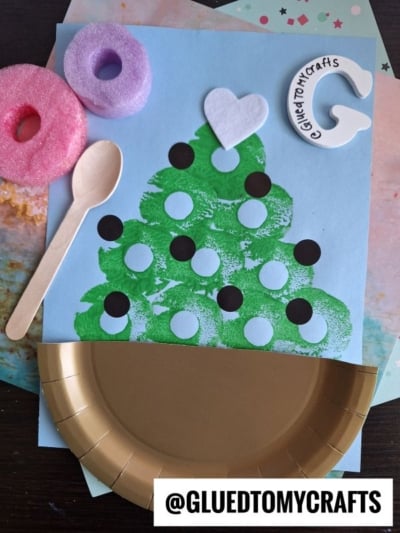 A handmade ice cream bowl craft using green paint, black dots, a paper heart, a craft stick, and a gold paper plate. Pool noodle crafts supplies like pool noodles and even a wooden spoon add fun touches. @GLUEDTOMYCRAFTS is written at the bottom.