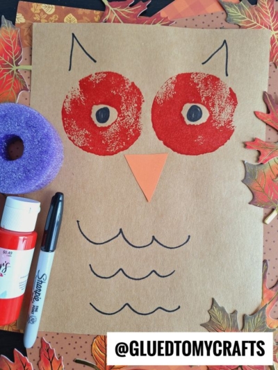 A brown paper with a stamped red owl face, drawn eyes, orange beak, and black wings—perfect for pool noodle crafts. Surrounded by fall leaves, purple sponge, glue stick, and marker. @GLUETOMYCRAFTS label at the bottom.