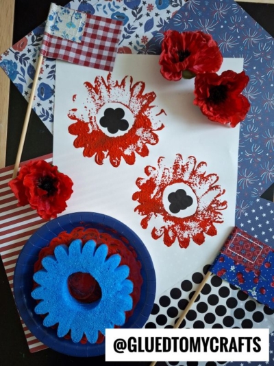 Patriotic-themed poppy flower art made with red and black paint on white paper, surrounded by artificial red poppies, small American flags, patterned blue and red paper, a blue foam flower shape, and a creative pool noodle craft. @GLUEDTOMYCRAFTS is written at the bottom.