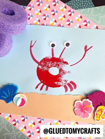 A kids’ craft of a red crab made with a painted pool noodle craft donut shape and googly eyes on blue paper, decorated with foam shapes of a shell, beach ball, and flower. The handle @gluedtomycrafts is at the bottom.