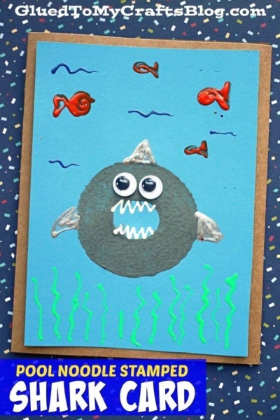 A handmade card features a shark made from a gray pool noodle craft stamp, with googly eyes and drawn fins and teeth. Red fish and green seaweed decorate the blue background. Text reads: POOL NOODLE STAMPED SHARK CARD.