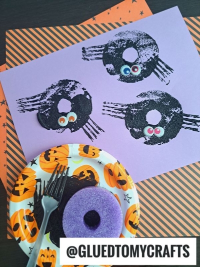 A Halloween craft scene with three black spider prints made from donuts on purple paper, each with googly eyes. A plate holds a purple donut and a fork, on top of pumpkin-themed paper—perfect inspiration for pool noodle crafts! Text reads @GLUEDTOMYCRAFTS.