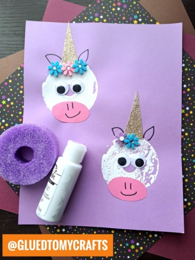 A purple paper displays two unicorn faces made from white paint prints—created with a pool noodle craft—plus googly eyes, pink snouts, gold glitter paper horns, and blue flowers. A paint bottle and a purple stamper sit beside the artwork.
