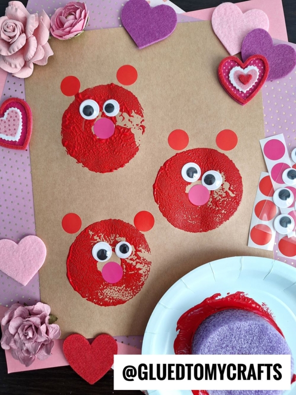 A craft project featuring three red bear faces with googly eyes and pink noses, made with paint and paper circles on brown paper, surrounded by felt hearts and artificial flowers—a charming take on pool noodle crafts. @GLUEDTOMYCRAFTS text is at the bottom.