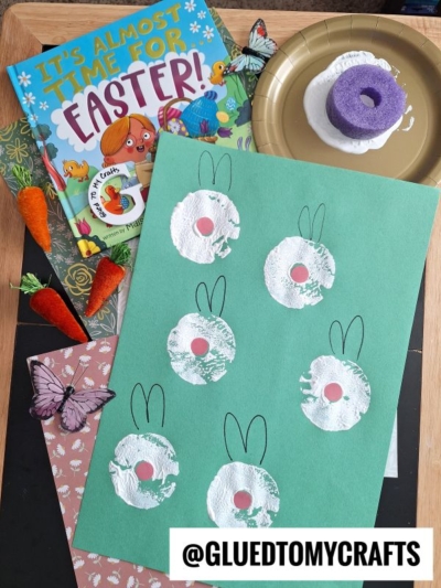 A green paper displays six white bunny prints made with a round sponge, perfect for pool noodle crafts. Nearby are a children’s Easter book, fake carrots, a purple sponge on paint, and a butterfly decoration.
