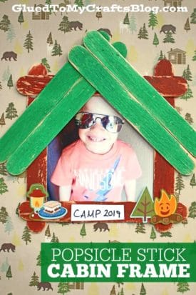A child smiling inside a popsicle stick cabin picture frame, radiating warmth and creativity.