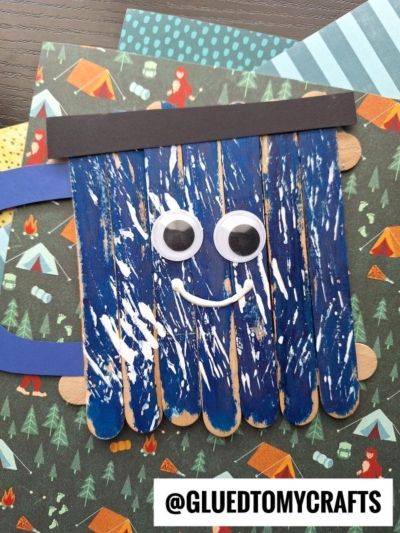 A popsicle stick camping mug craft, painted blue with a black paper strip on top, large googly eyes, and a white pipe cleaner smile. The background features camping-themed paper. The text @GLUEDTOMYCRAFTS is at the bottom.