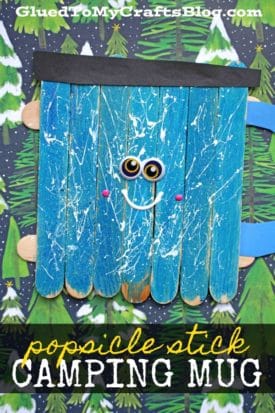 A popsicle stick camping mug with googly eyes, a smiling face, and a handle sits against a forest-patterned background. The blue mug features white paint splatters, a black strip at the top, and creative charm.