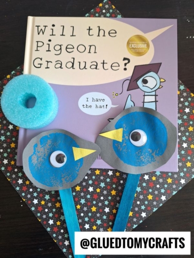 A children’s book titled Will the Pigeon Graduate? sits on a star-patterned background. In front are two blue pigeon pool noodle crafts with googly eyes and yellow beaks, plus a blue foam ring. Text at bottom: @GLUEDTOMYCRAFTS.