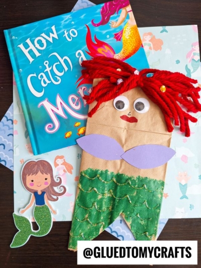 A children’s book titled “How to Catch a Mermaid,” a mermaid paper bag puppet with red yarn hair, googly eyes, green scales, fun mermaid paper party bags, a cartoon mermaid cutout, and the @GLUETOMYCRAFTS label.