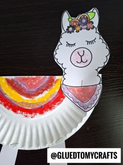A paper plate llama friend is decorated as a colorful llama with a drawn face, floral crown, and a patterned scarf. This fun project shows how to make a paper plate llama that’s both playful and creative. The bottom displays the @GLUEDTOMYCRAFTS watermark.