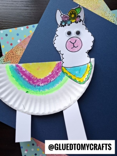A vibrant llama paper plate craft featuring a decorated head, googly eyes, and drawn details, set on blue and polka dot paper. The handle @GLUEDTOMYCRAFTS is displayed at the bottom.