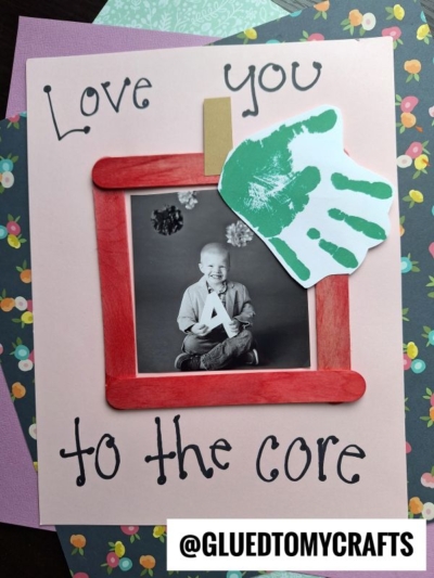 A handmade card featuring the phrase “Love you to the core,” a photo of a smiling child, and a charming craft stick apple picture frame with a red border and green handprint cutout. The @gluedtomycrafts handle is displayed at the bottom.