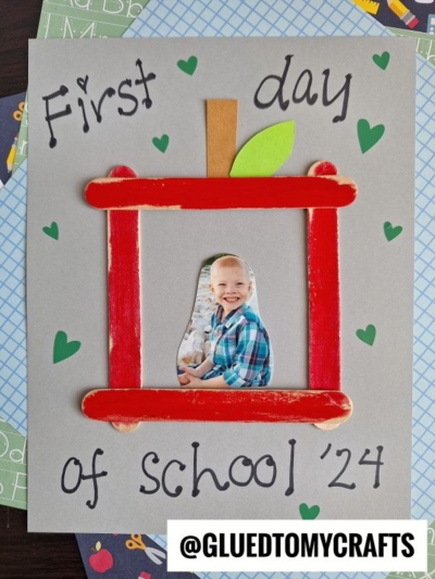 A handmade First Day of School 24 card featuring a craft stick apple picture frame around a child’s photo. Small green hearts decorate the gray background.