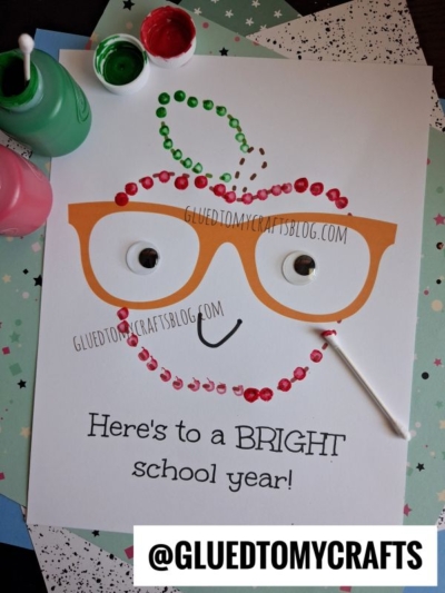 A cheerful paper craft of a q-tip painted back to school apple with orange glasses, googly eyes, and a wide smile, made using colorful paint dots. The text reads, "Heres to a BRIGHT school year!" Nearby are paint pots, a cotton swab (q-tip), and a brush—everything you need for your own creative q-tip painted apple.