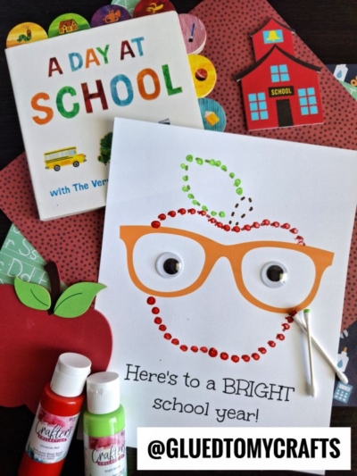 A school-themed flat lay features A Day at School book, a red paper apple, paint bottles, and a q-tip painted back to school apple craft with orange glasses and googly eyes, plus “Here’s to a BRIGHT school year!” written below.