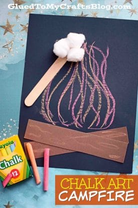 A chalk art campfire craft on black paper features colorful chalk flames, two brown paper strips for logs, a craft stick, and three cotton balls at the top. Crayola chalk and extra pieces lie beside this vibrant paper art campfire.