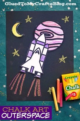 A colorful chalk art outer space drawing of a rocket ship blasting off, with a crescent moon, stars, and striped planet in the background. Crayola chalk sticks and a chalk box make this paper chalk art outer space scene come alive.