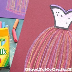 A purple construction paper showcases a chalk art dress outline; the top is a paper cutout, while the skirt, colored with pink and orange chalk, pops vibrantly. A box of Crayola chalk sits nearby on the textured blue background.