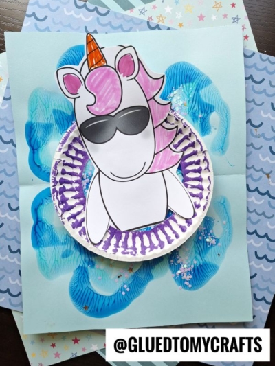 A pool float unicorn craft with pink hair and sunglasses, made from paper and a painted paper plate, is placed on blue ocean-themed paper. The username @GLUEDTOMYCRAFTS is displayed at the bottom.