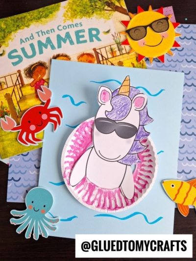 A summer-themed craft scene with a pool float unicorn craft wearing sunglasses in a cupcake liner, surrounded by cutouts of a sun, crab, octopus, and fish, with the children’s book And Then Comes Summer in the background.
