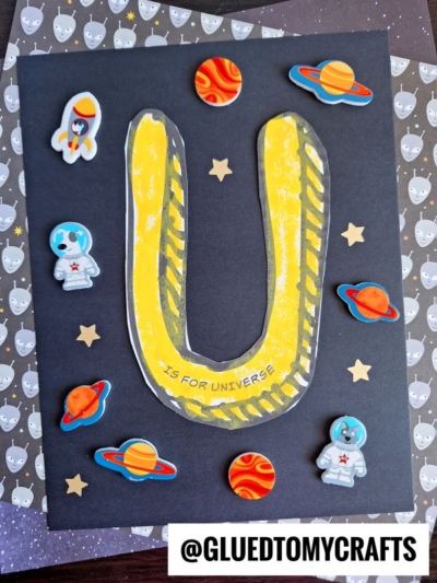A black card features a large, yellow letter U labeled is for Universe. This playful paper universe craft includes cartoon planets, astronauts, rockets, and star stickers on a cat-patterned background. @GLUEDTOMYCRAFTS is at the bottom.