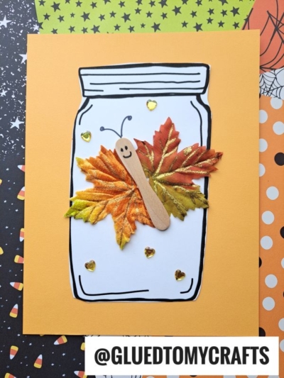 A paper craft of a jar on orange paper features a leaf butterfly in jar, made from colored leaves and a popsicle stick with a smiley face. Gold heart stickers decorate the jar, and the background has Halloween-themed patterns.