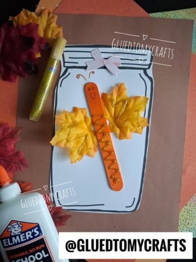 A craft featuring a mason jar outline with a smiling dragonfly made from a popsicle stick, fake leaves for wings—creating the look of a whimsical leaf butterfly in jar—and sparkling gold glitter. Elmer’s glue, gold glitter glue, and fake leaves are nearby.