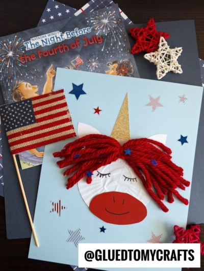 A handmade cupcake liner unicorn craft with red yarn hair, a gold horn, and a paper American flag sits on a light blue background, surrounded by patriotic-themed decorations and the book The Night Before the Fourth of July.