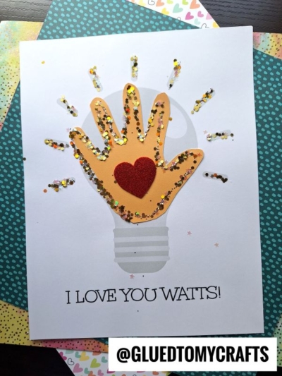 A handmade card features a handprint light bulb with a red heart in the center, decorated with glitter to resemble a glowing effect. Below, the text reads, I LOVE YOU WATTS! with the handle @GLUEDTOMYCRAFTS.