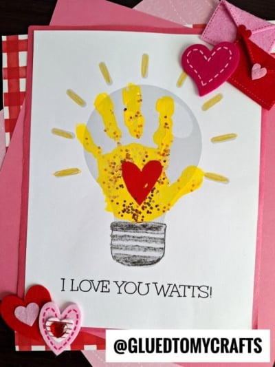 A handprint light bulb painted yellow, sprinkled with glitter and topped with a red heart, is featured on a card reading I LOVE YOU WATTS! and surrounded by felt hearts and Valentine-themed decorations.