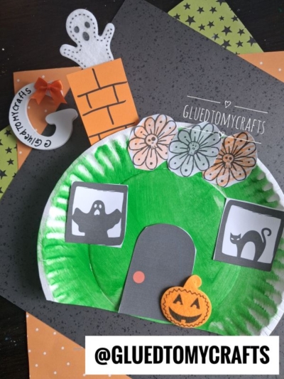 A Halloween-themed paper plate craft features ghosts, a black cat, flowers, and a pumpkin—inviting you to create your own spooky scene reminiscent of a paper plate haunted house. Craft supplies and orange paper are displayed underneath. @GLUEDTOMYCRAFTS is written across the bottom.