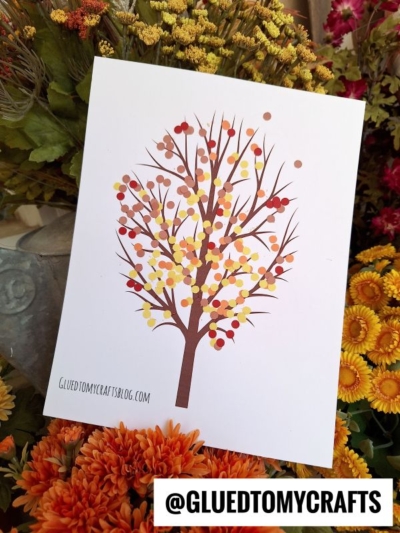 A fall-themed art print of a tree with multicolored dot leaves in red, yellow, and orange tones—resembling paper punch confetti—placed among vibrant orange, yellow, and green flowers. @GLUEDTOMYCRAFTS is displayed at the bottom of the image.