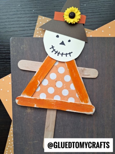 Create a charming popsicle stick scarecrow crafted from painted popsicle sticks, decorated with polka-dot paper, a paper hat adorned with a sunflower, and a drawn-on smiling face. These delightful popsicle stick scarecrow puppets are displayed on a dark surface with orange dotted paper accents, adding a playful touch to your seasonal decor.